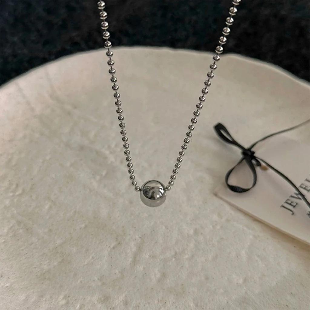 Collier à pendentif bille posé à plat sur une coupelle claire, chaîne à billes en acier argenté.