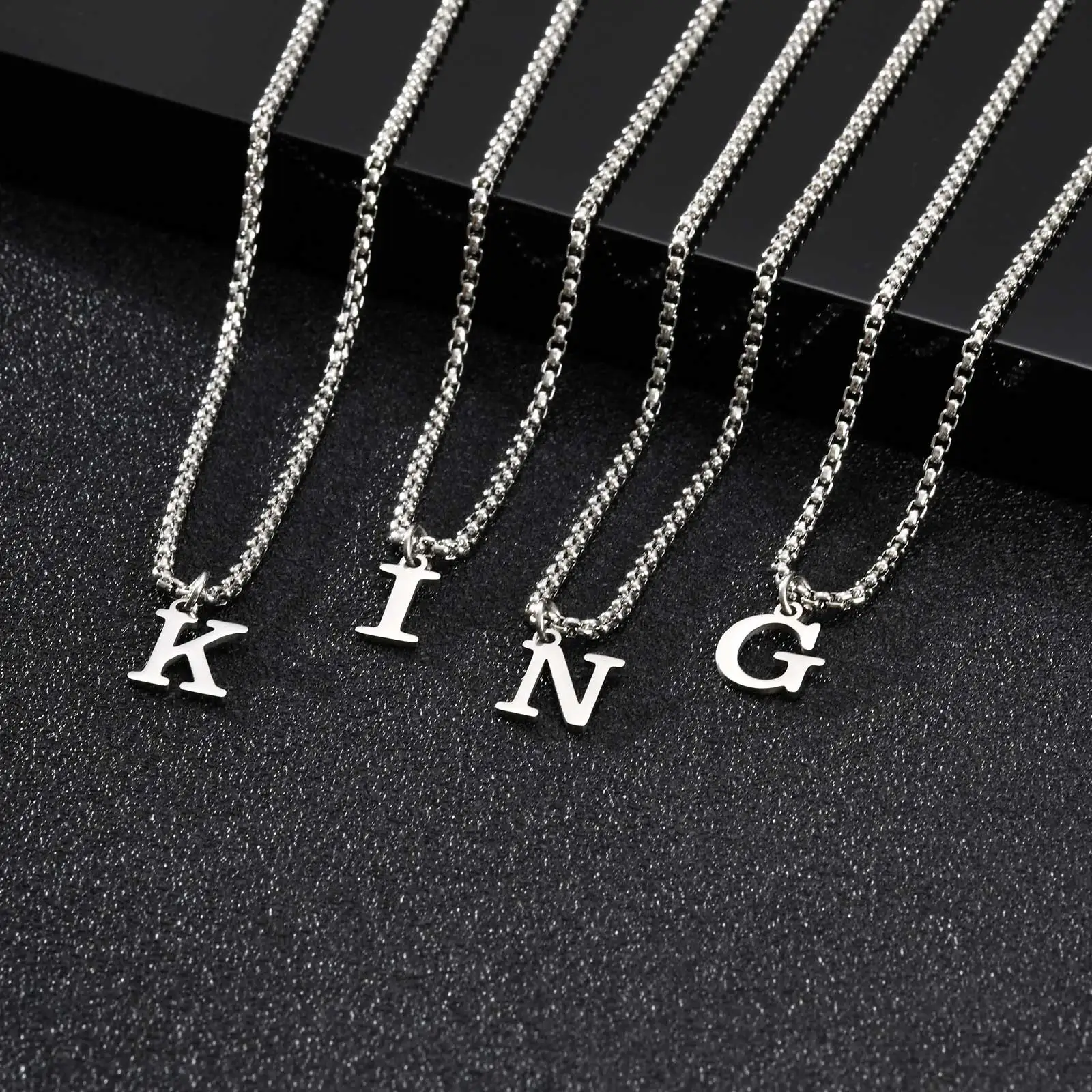 Quatre pendentifs lettres K, I, N et G en acier poli, alignés sur leurs chaînes maille boîte argentées
