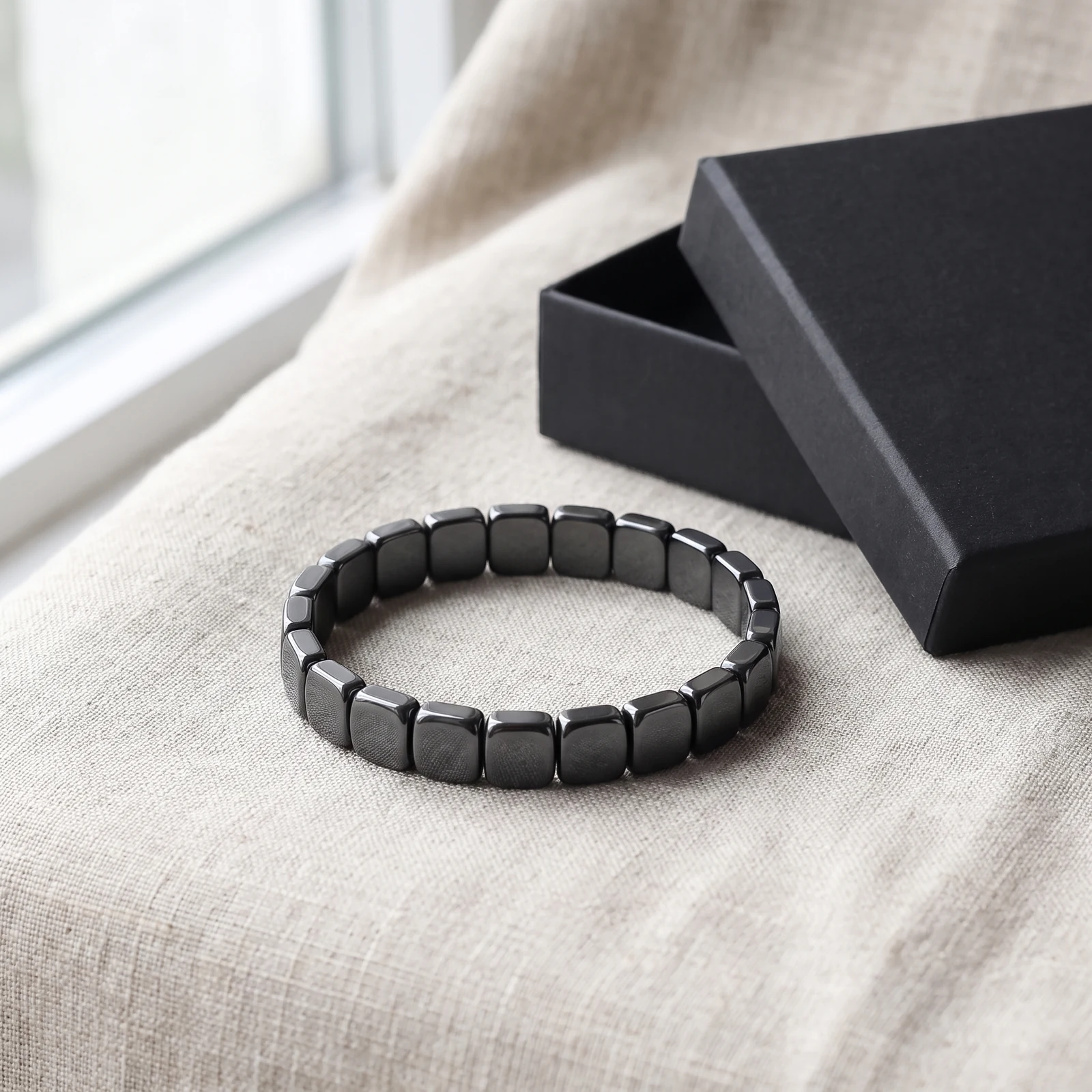 Bracelet homme en perles d'hématite présenté sur du lin beige à côté d'un coffret cadeau noir